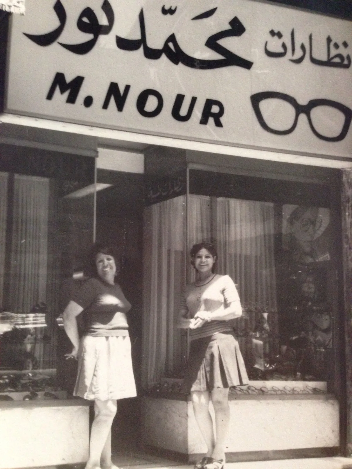 Black and white photo from 1960 of two women smiling outside the M. Nour optical shop in Alexandria. The sign reads M. Nour in English and Arabic, next to a drawing of a pair of glasses.